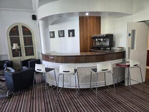 A hotel lobby with a curved reception desk, bar stools, a coffee machine, and a patterned carpet.