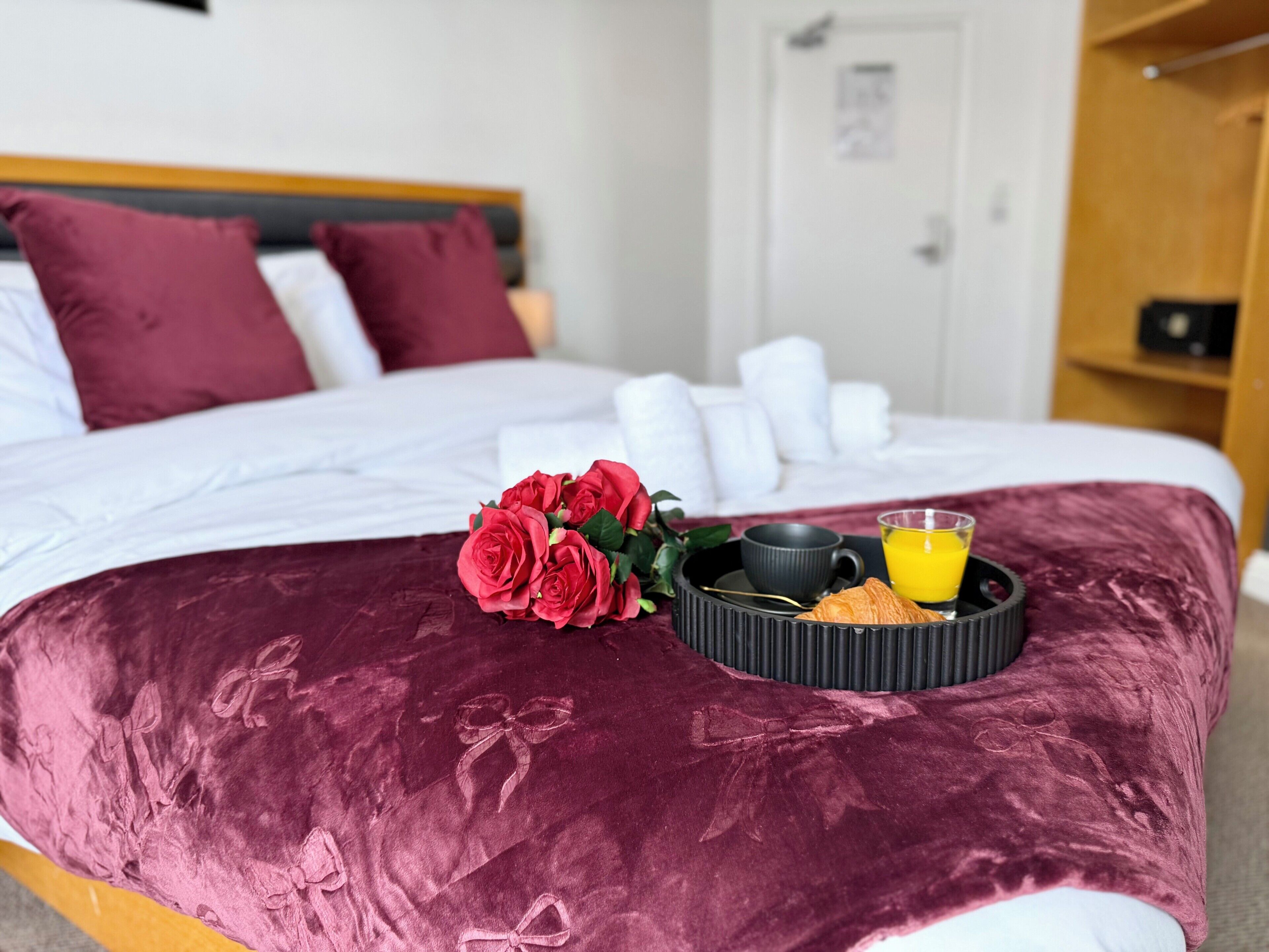 A bed with a dark red bedspread, a bouquet of red roses, a black tray with a cup and a glass of orange juice, and a wooden headboard.