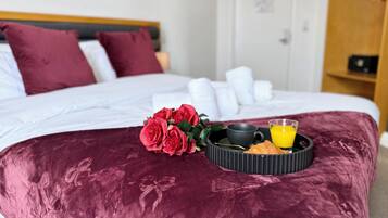 A bed with a dark red bedspread, a bouquet of red roses, a black tray with a cup and a glass of orange juice, and a wooden headboard.