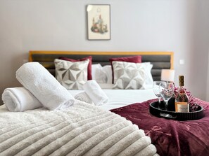 Habitación de hotel con una cama bien hecha, una botella de vino y una bandeja con vasos sobre un edredón de terciopelo rojo.