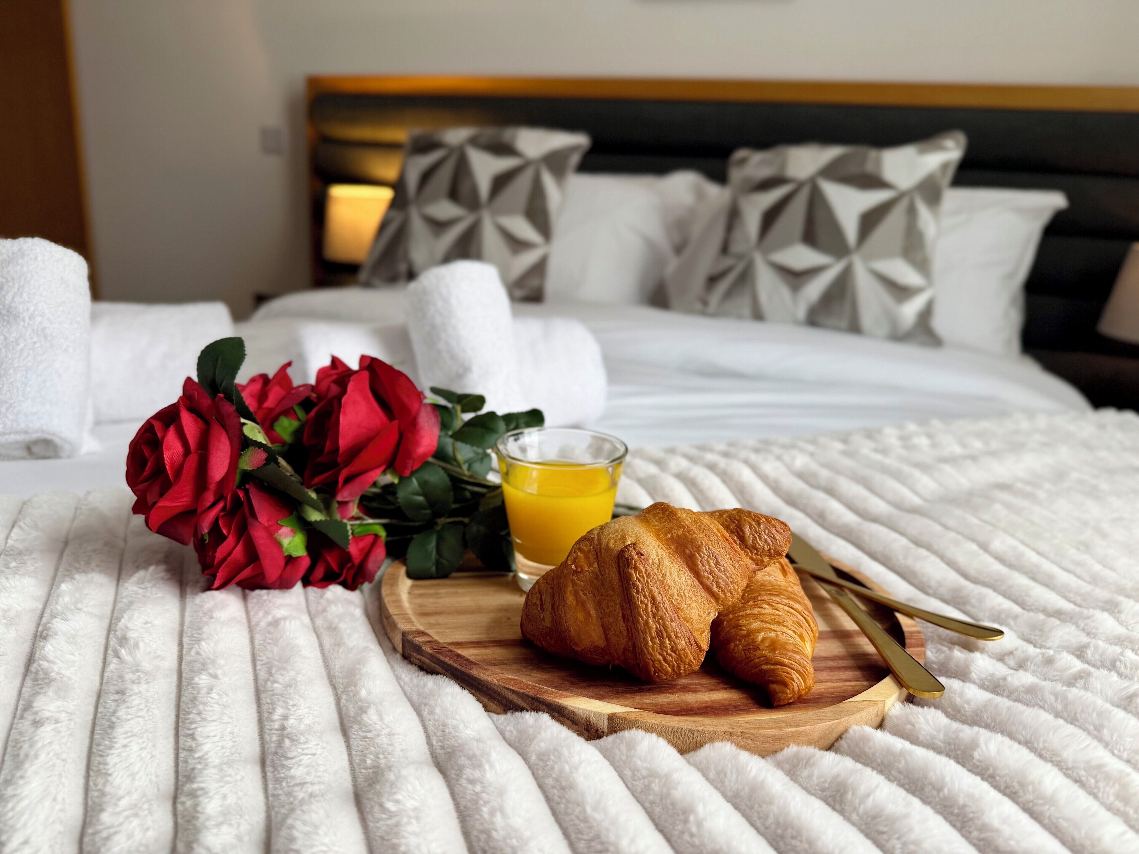 Una cama con edredón blanco, una bandeja de madera con croissants y un vaso de zumo de naranja, y un ramo de rosas rojas.