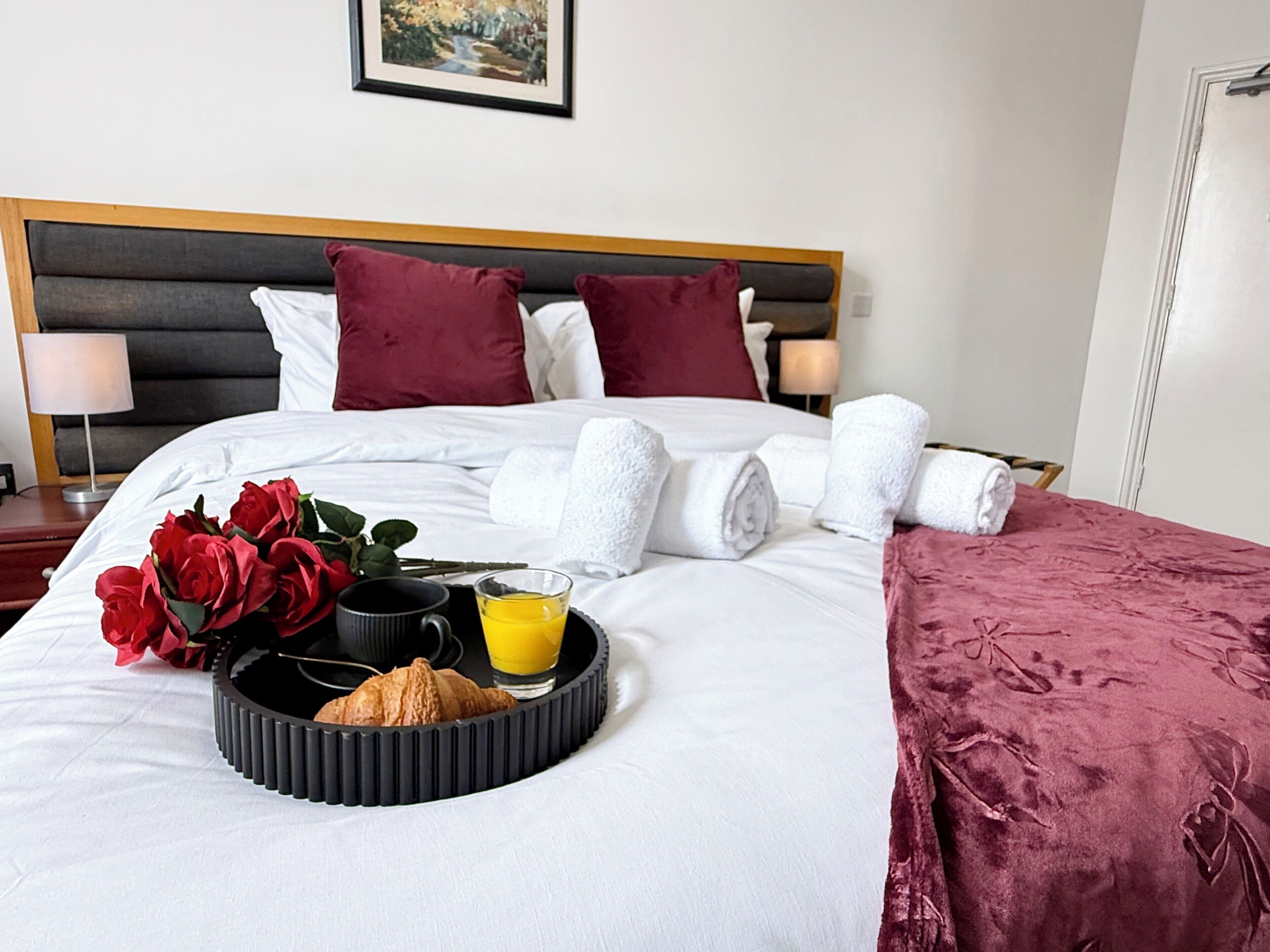 A neatly made bed with white linens, a tray with croissants and orange juice, and a bouquet of red roses.