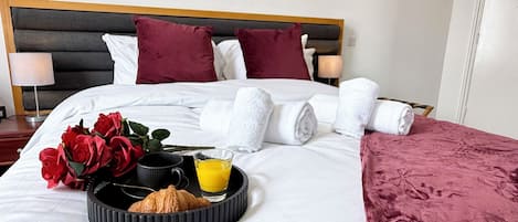A neatly made bed with white linens, a tray with croissants and orange juice, and a bouquet of red roses.
