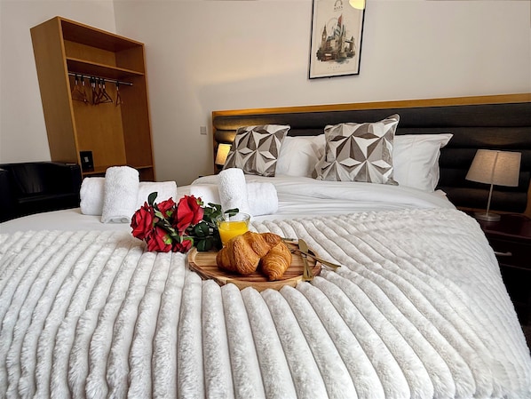 Una cama bien hecha con un edredón blanco mullido, una bandeja con cruasanes y un vaso de jugo de naranja, y un ramo de rosas rojas.