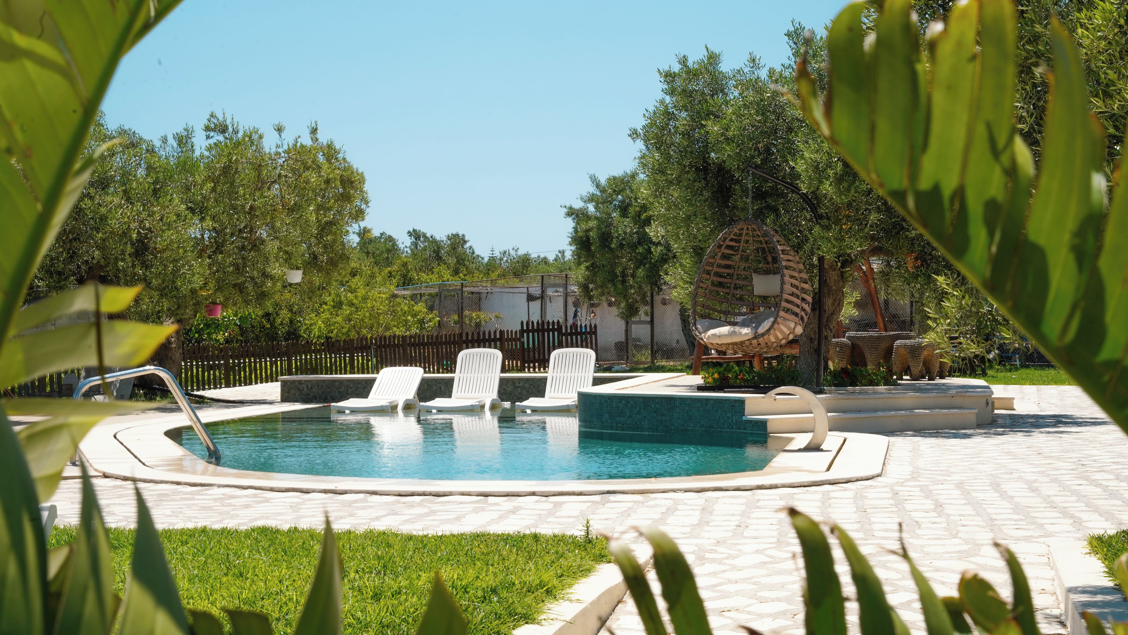 Piscina con sillones de descanso y una hamaca colgante, rodeada de vegetación y una valla.