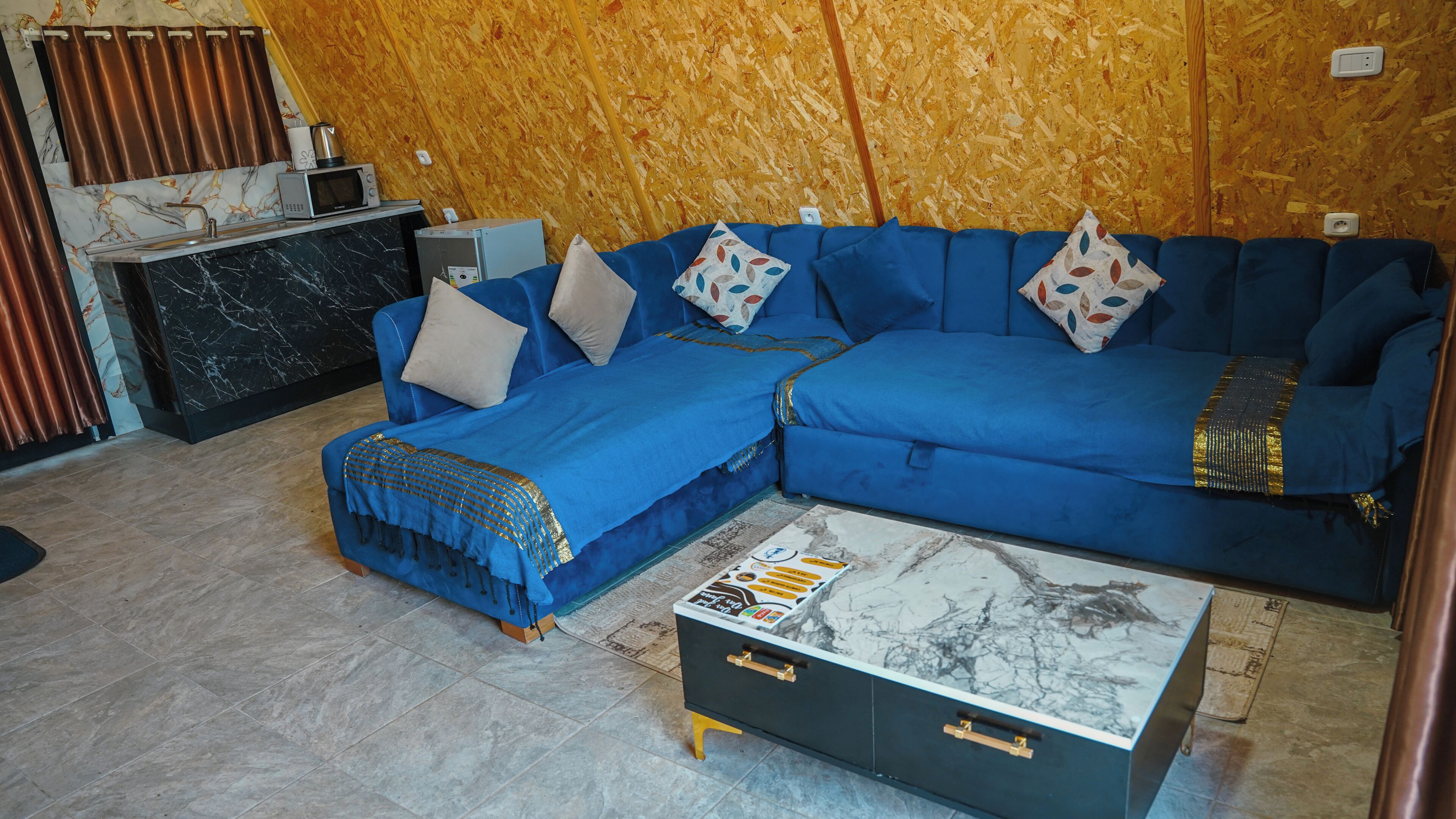 A room with a blue sectional sofa, a marble coffee table, a small cabinet, and a wooden wall.
