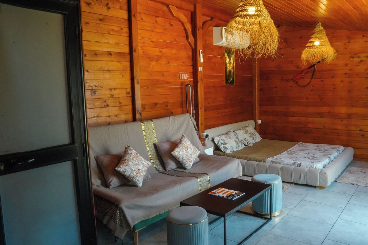 Interior de una cabaña de madera con sofá, dos camas individuales, una mesita y lámparas colgantes.