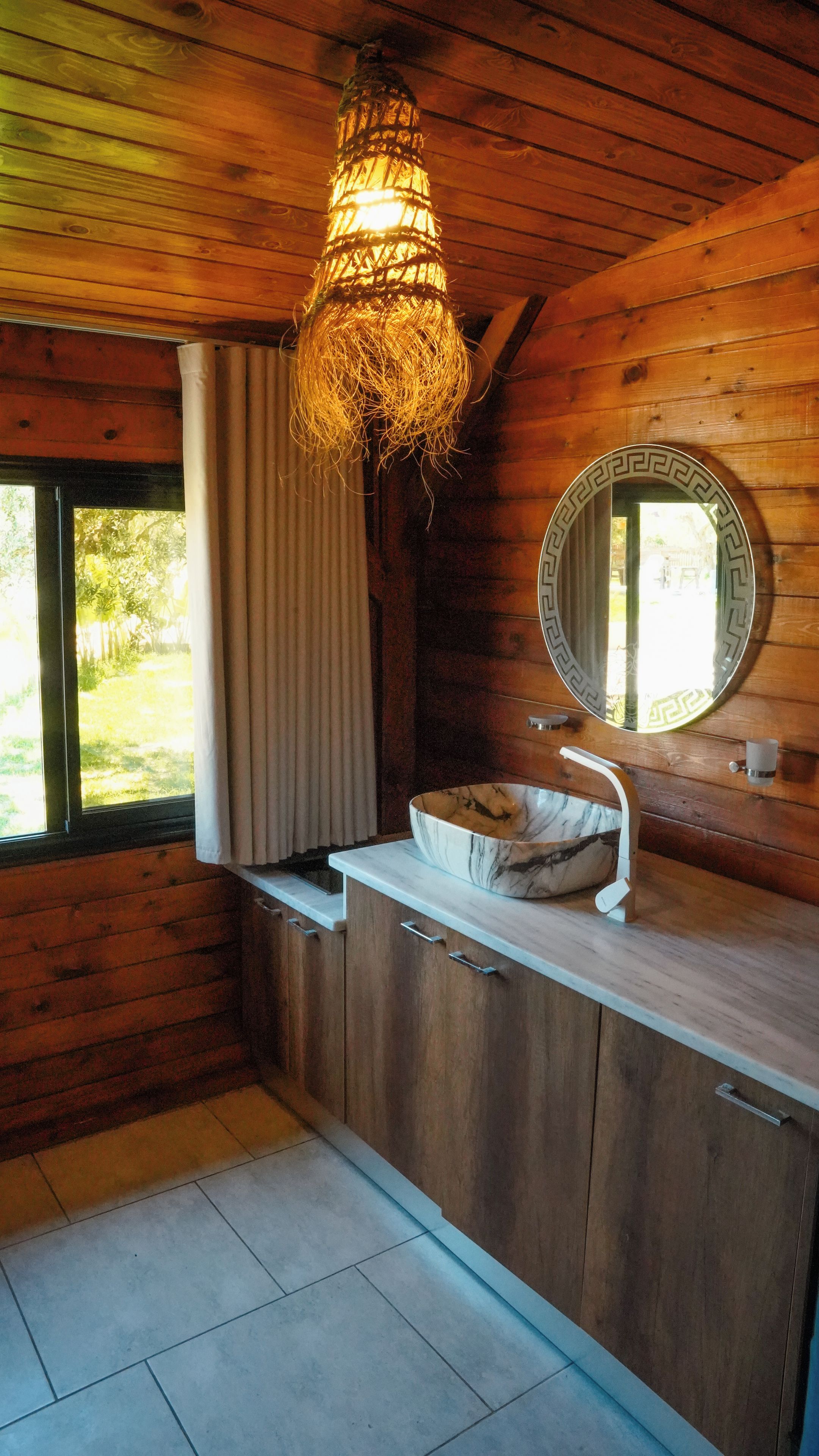 L’intérieur d’une cabane en bois, avec un évier encastré, un luminaire suspendu et une fenêtre avec des rideaux.