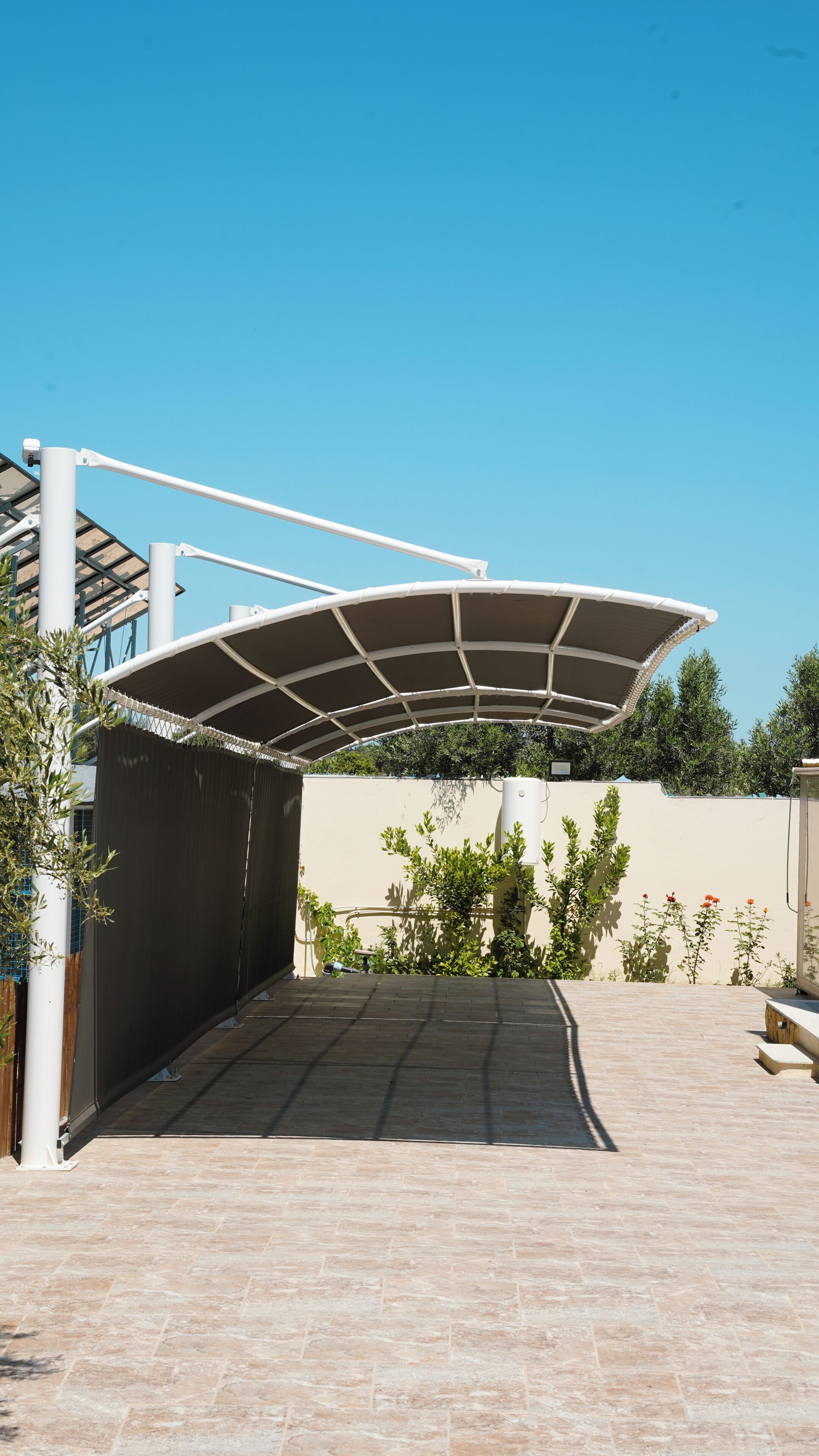 A covered outdoor area with a tiled floor, a white structure with a curved roof, and a beige wall with greenery.