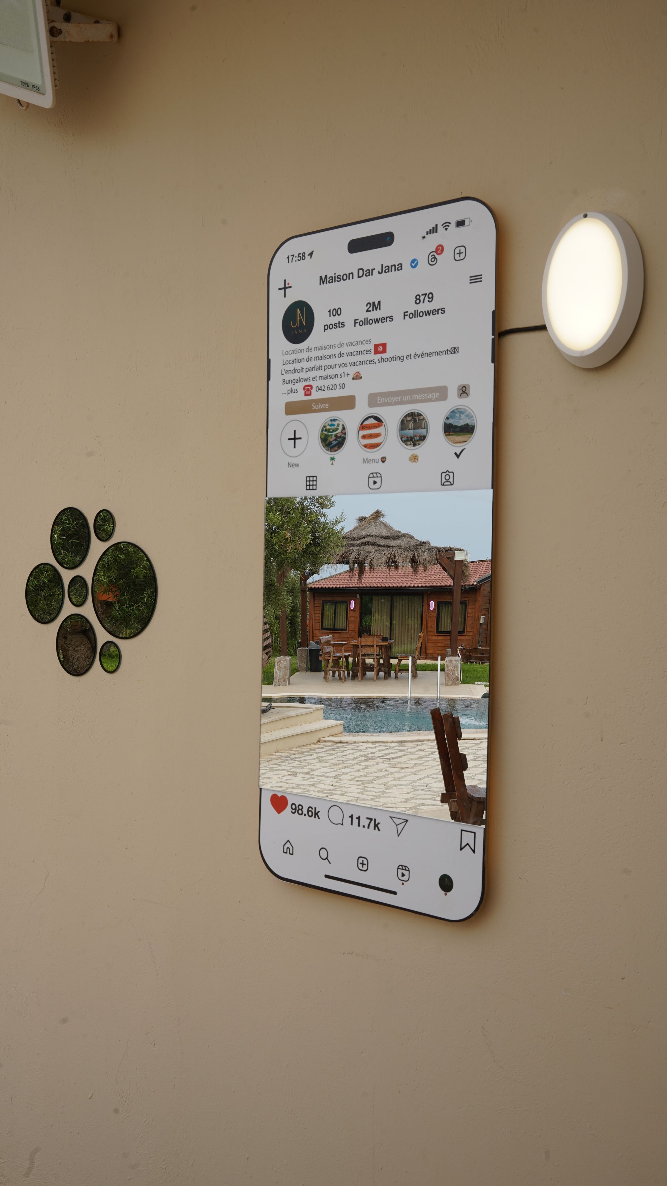 En smartphone viser en annonce for en feriebolig med pool, træmøbler og et stråtækt hus.
