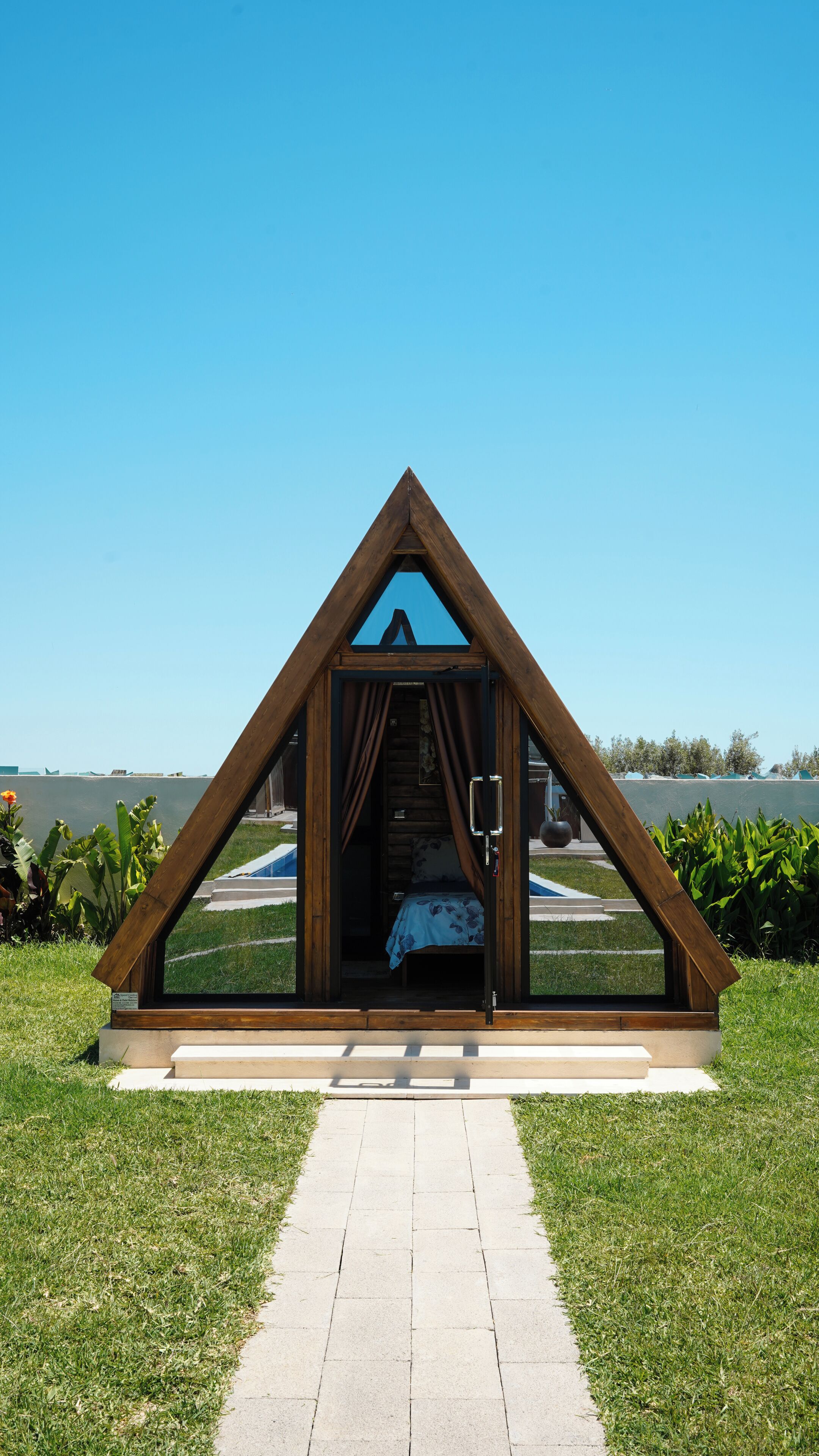 A triangular wooden structure with a bed inside, a clear sky, and a pathway leading to it.