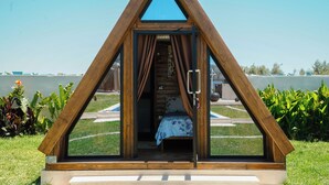A triangular wooden structure with a bed inside, a window, and a view of a pool and garden. - Dar Jana et Dar Jad (Ben Khiar Diar Ben Salem)