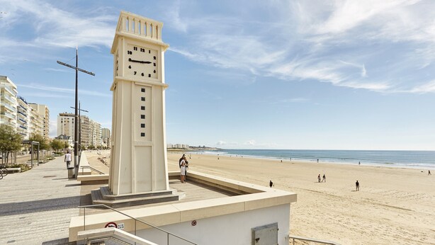 Ein Glockenturm an einer Promenade mit Blick auf einen Strand, im Hintergrund sind Gebäude zu sehen.