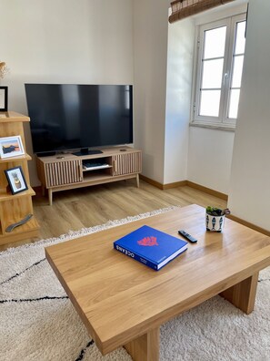 En stue med et fladskærms-tv på et træstel, et sofabord med en bog og fjernbetjening, en lille potteplante og en hvid måtte.