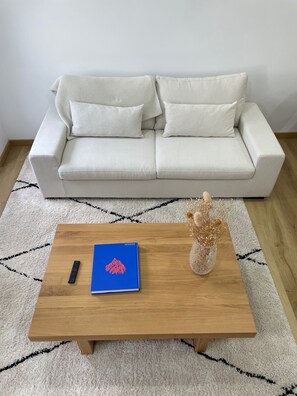 Ein Wohnzimmer mit einem weißen Sofa, einem Holztisch, einer Fernbedienung, einem blauen Buch und einer dekorativen Vase.