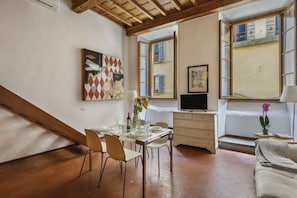 A dining area with a table set for four, a television on a stand, and a window with shutters. - Via Maggio 10 – Historic Loft in Oltrarno  (Florence)