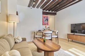 Ein modernes Wohnzimmer mit einer Couch, einem Couchtisch, einer Essecke mit Stühlen, einem Fernseher und einer Decke mit sichtbaren Balken.