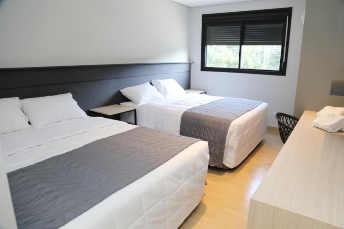 Habitación de hotel con dos camas, una ventana con persianas, una mesita de noche y una pequeña zona de escritorio.