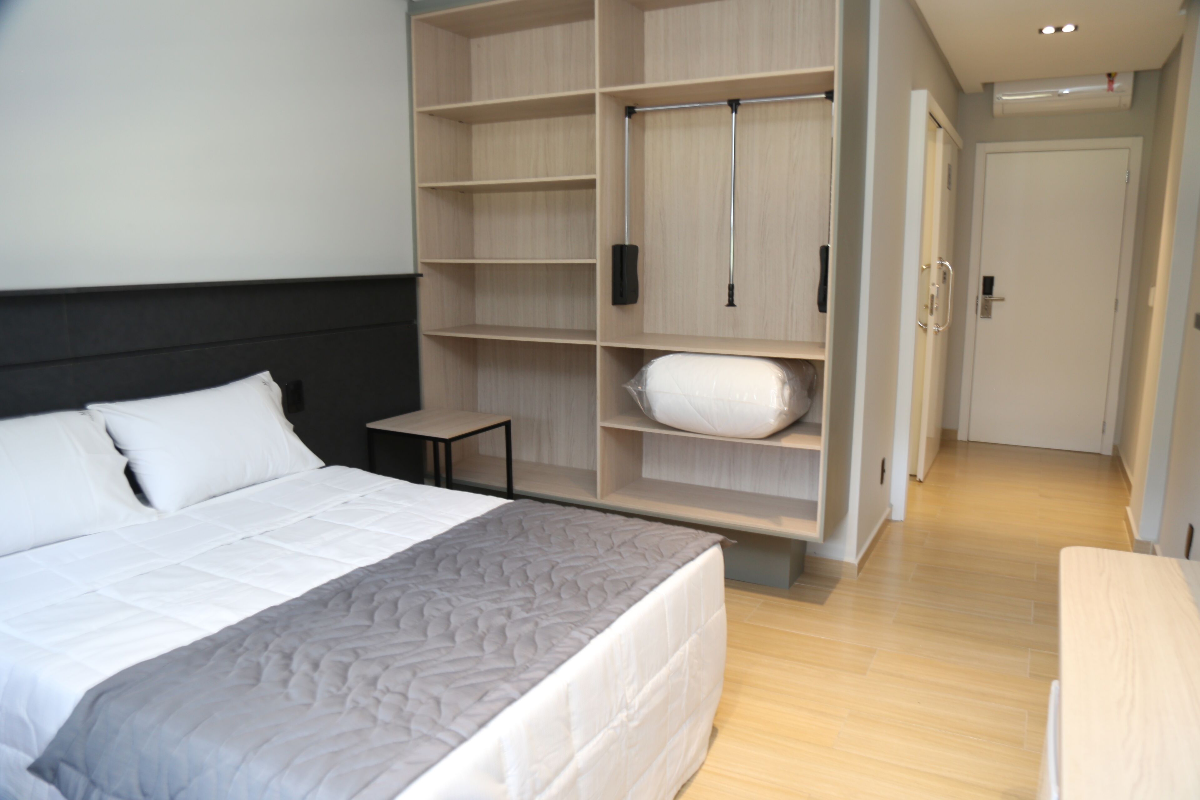 A hotel room with a large bed, a wardrobe with multiple shelves, a small bedside table, and a door leading to another room.