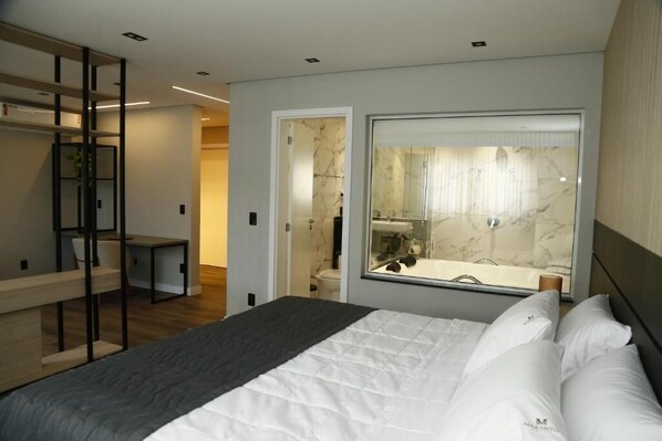 A modern bedroom with a bed, a desk, and a bathroom visible through a glass door.