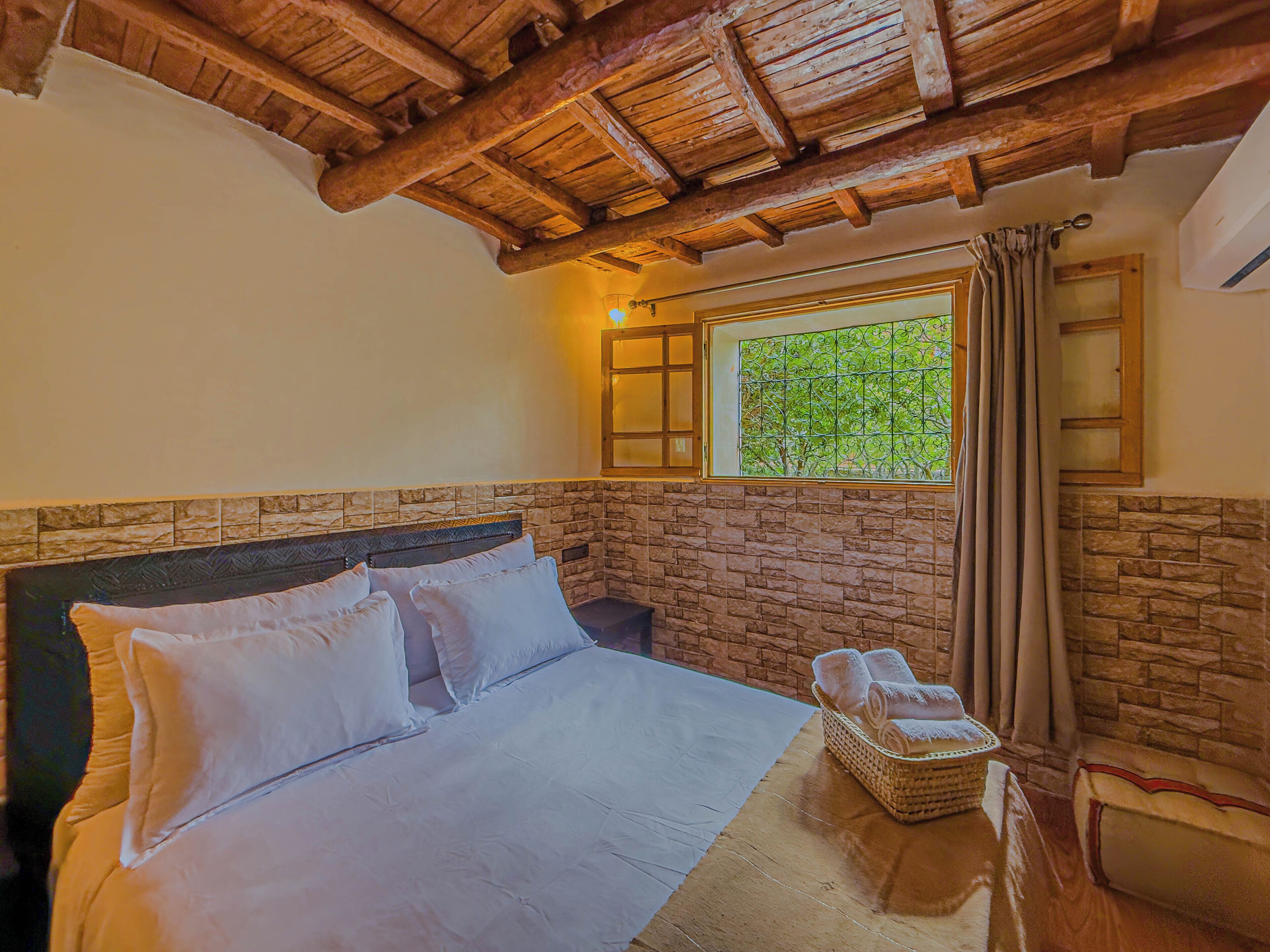 Une chambre avec un mur en pierre, un plafond en bois, un lit avec du linge de maison blanc, une fenêtre donnant sur un beau paysage et un panier rempli de serviettes.
