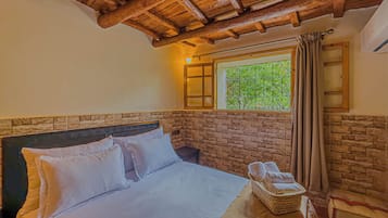 Une chambre avec un mur en pierre, un plafond en bois, un lit avec du linge de maison blanc, une fenêtre donnant sur un beau paysage et un panier rempli de serviettes.