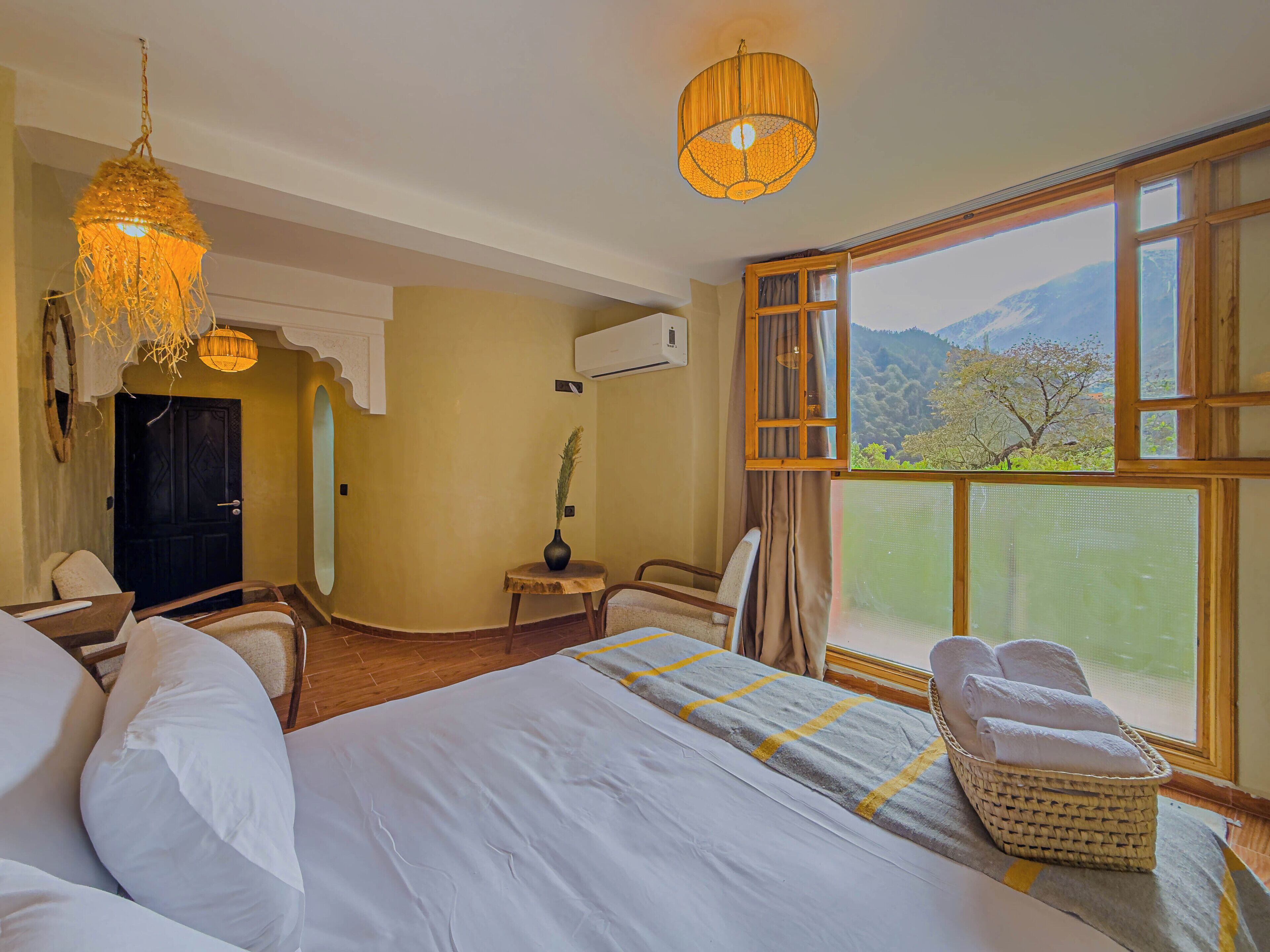 Habitación de hotel con una cama grande, una zona de estar con una silla y una mesita pequeña, una lámpara colgante decorativa y un ventanal con vistas al exterior.