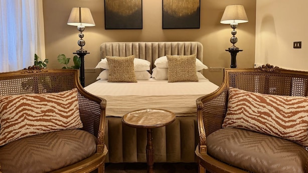 Quarto de hotel com cama, duas cadeiras, uma mesinha e duas luminárias.