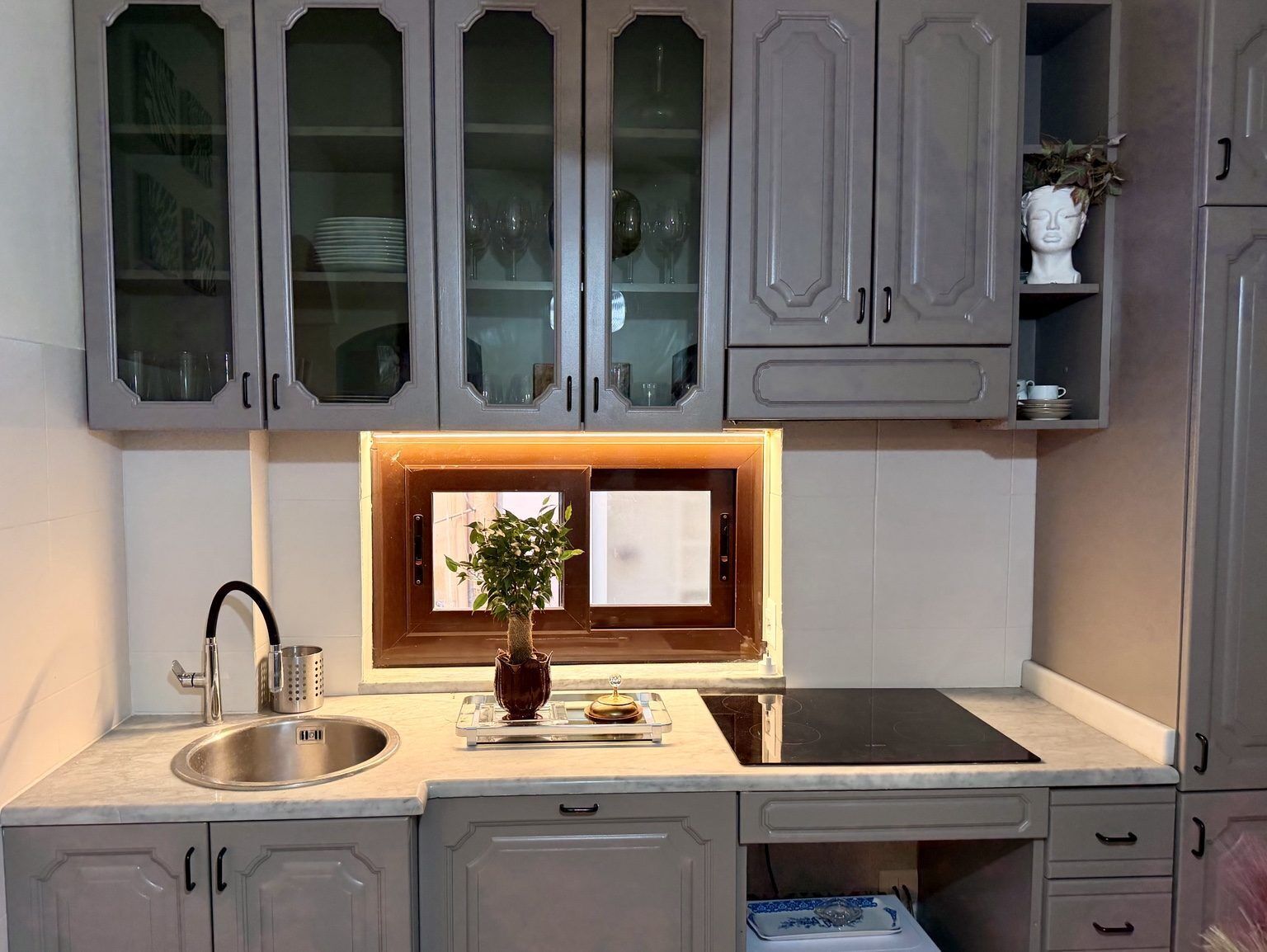 A kitchen with grey cabinets, a stainless steel sink, a built-in stove, and a window with a plant on the counter.