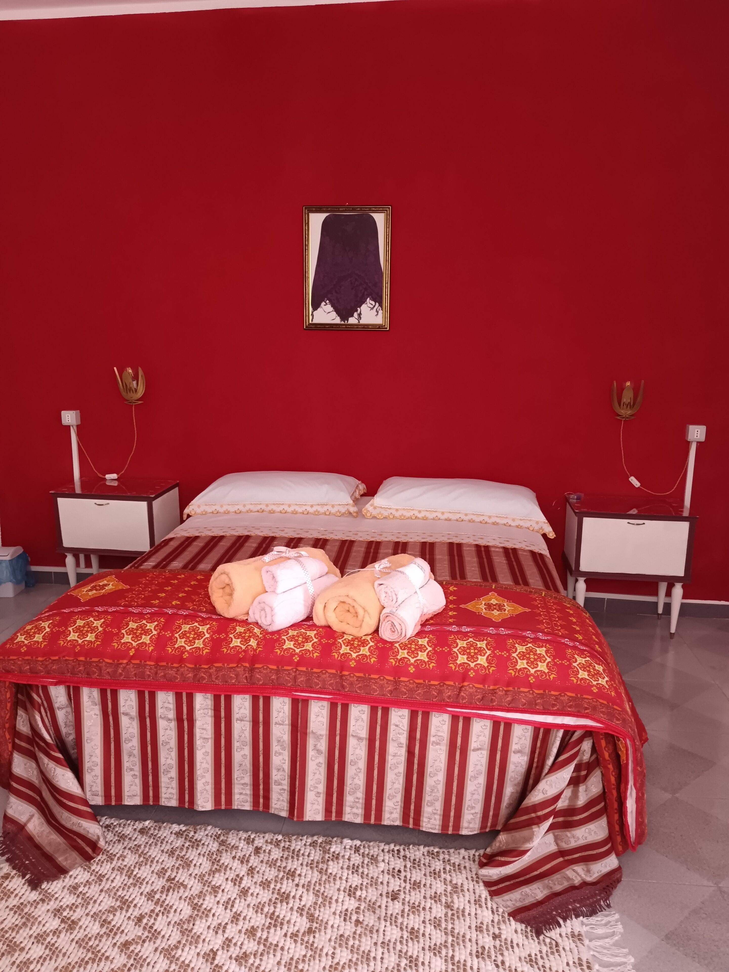 Habitación de hotel con pared roja, una cama con ropa de cama a rayas, dos mesitas de noche con lámparas y un cuadro enmarcado en la pared.