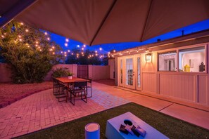 Ein Terrassenbereich mit Tisch und Stühlen, eine überdachte Sitzgelegenheit sowie ein Gebäude mit Fenstern und Türen.