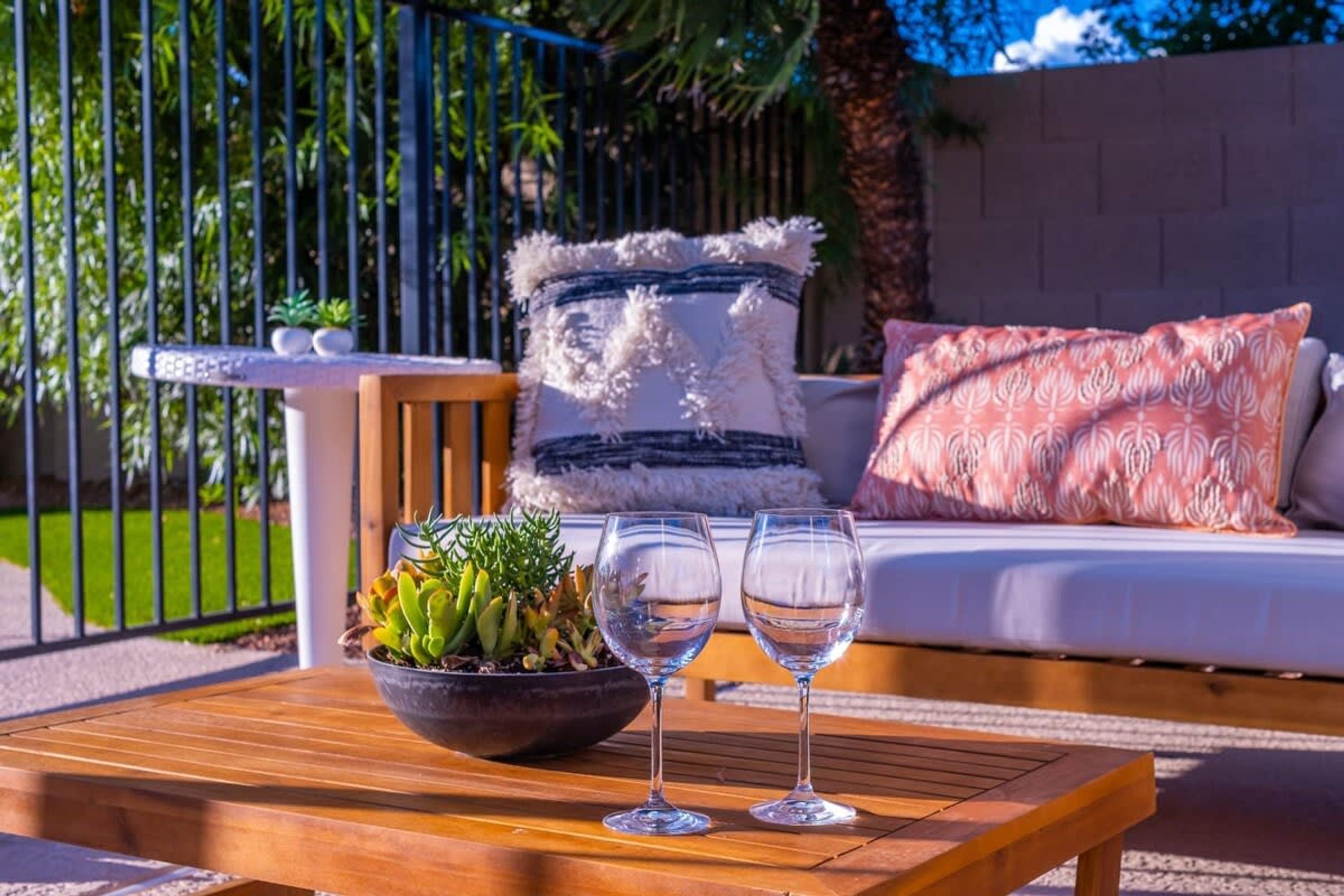 Uma mesa de madeira com uma tigela de plantas e dois copos de água, um sofá branco com almofadas estampadas, e uma grade de metal ao fundo.
