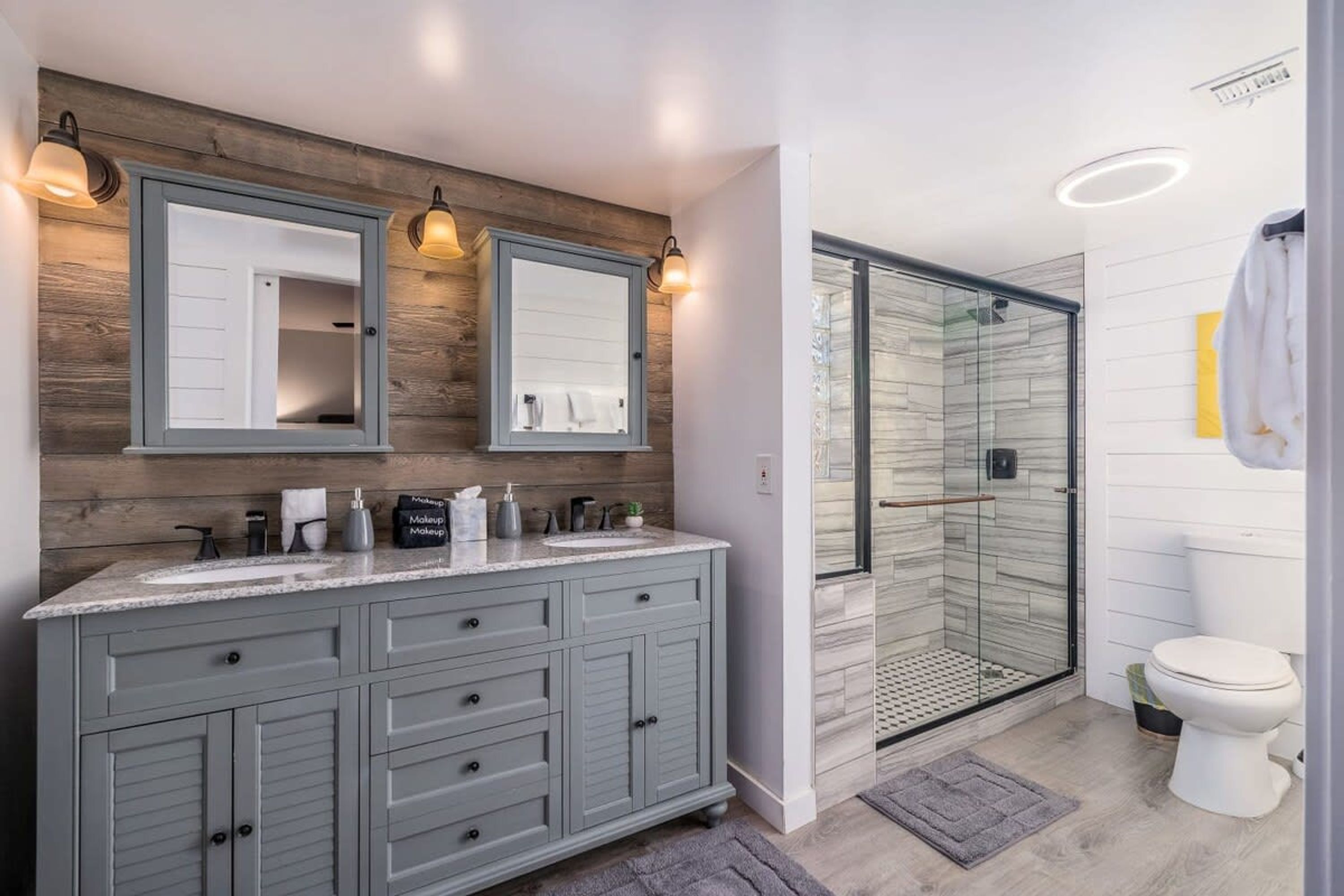 A modern bathroom with a grey vanity, two mirrors, a glass shower enclosure, and a white toilet.