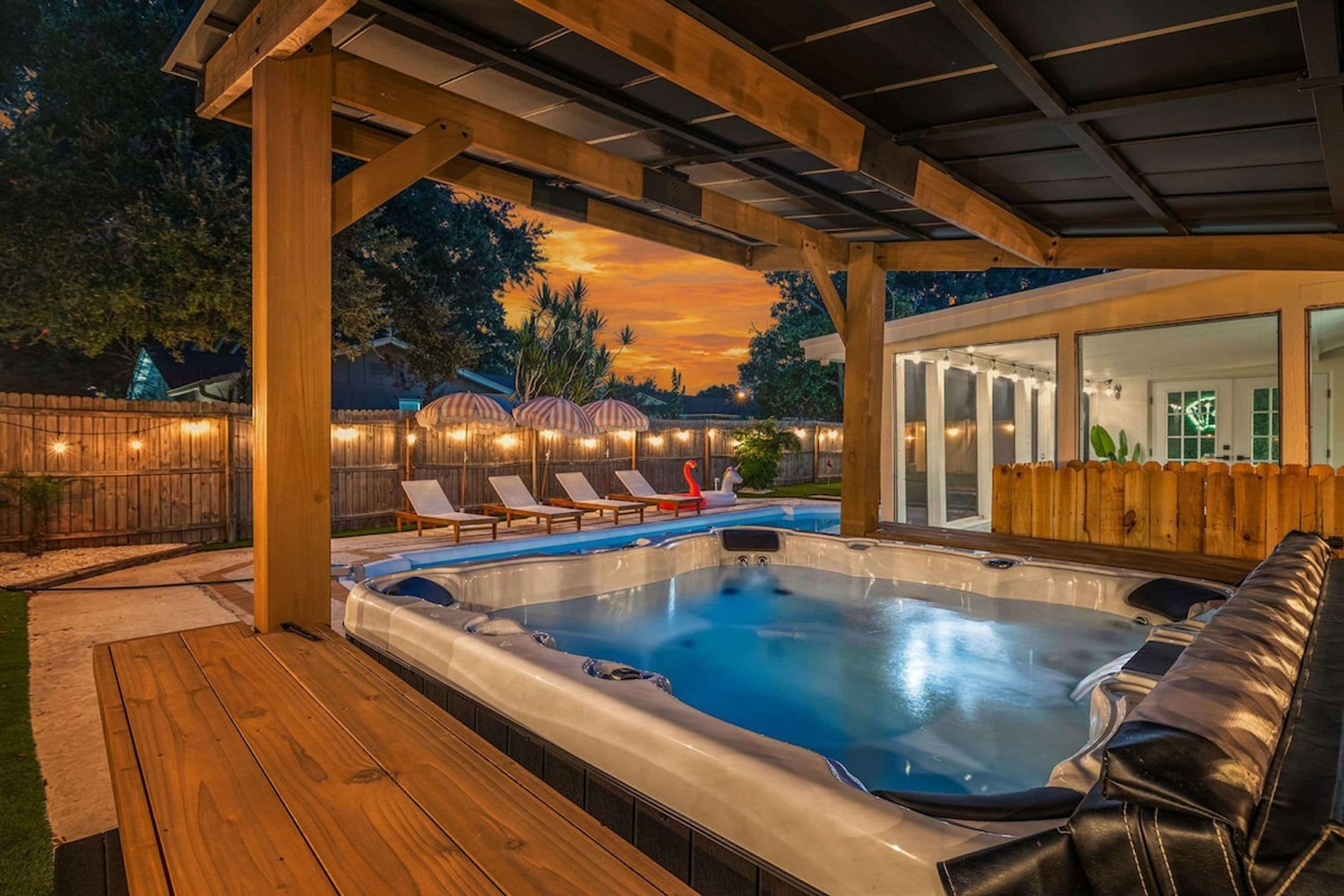 A hot tub under a wooden pergola with a view of a house and a sunset.