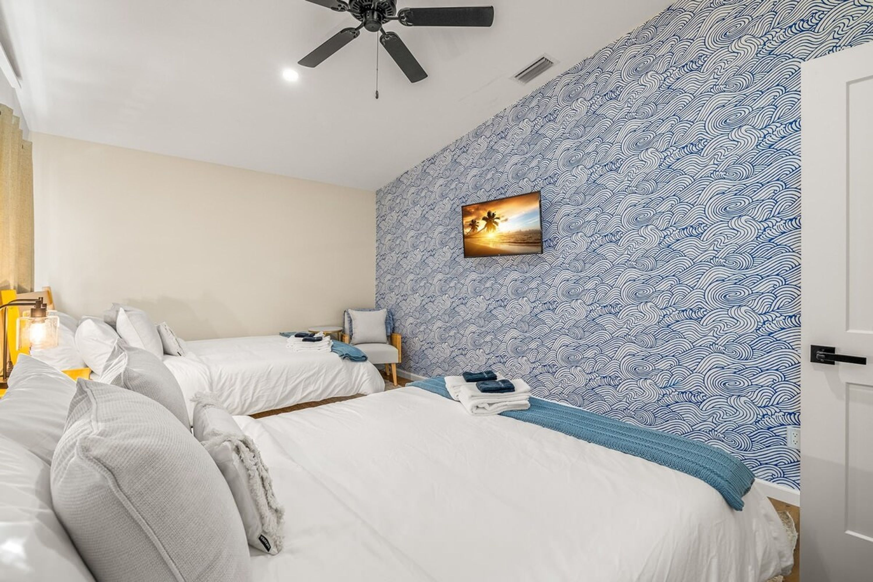 Une chambre d’hôtel avec deux lits, un ventilateur de plafond, un tableau accroché au mur et un papier peint à motifs.