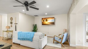 A modern living room with a ceiling fan, a flat-screen TV mounted on the wall, a white sofa with a blue throw, a wooden coffee table, and a side chair. - Beach Bliss Movie Theater Putt Putt Pool (Largo)