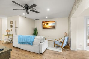 Ein modernes Wohnzimmer mit Deckenventilator, einem an der Wand befestigten Flachbildfernseher, einem weißen Sofa mit blauer Tagesdecke, einem Holztisch und einem Sessel.