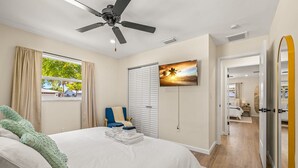 A bedroom with a bed, a ceiling fan, a window with curtains, a chair, a TV, and a door leading to another room. - Beach Bliss Movie Theater Putt Putt Pool (Largo)
