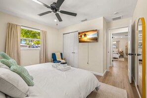 Ein Schlafzimmer mit einem Bett, einem Deckenventilator, einem Fenster mit Vorhängen, einem Stuhl, einem Fernseher und einer Tür zu einem anderen Raum.