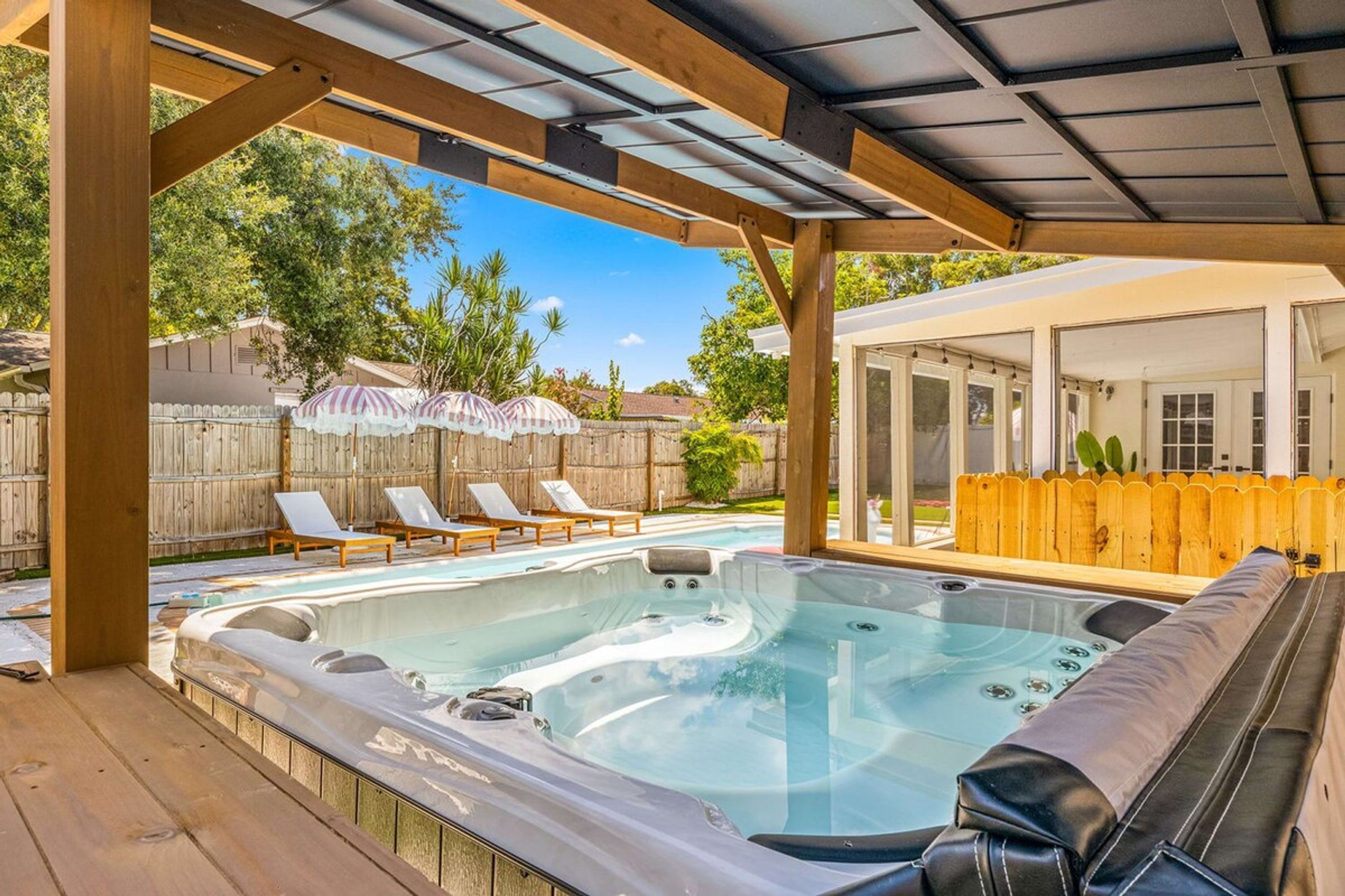 A hot tub under a wooden pergola with lounge chairs and umbrellas, a wooden fence, and a house in the background.