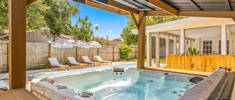 A hot tub under a wooden pergola with lounge chairs and umbrellas, a wooden fence, and a house in the background.