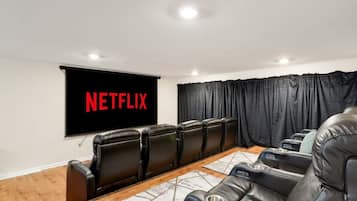Une salle de cinéma privée équipée d’un grand écran diffusant du contenu « NETFLIX », de fauteuils en cuir inclinables, d’une table basse en bois et d’un rideau noir.
