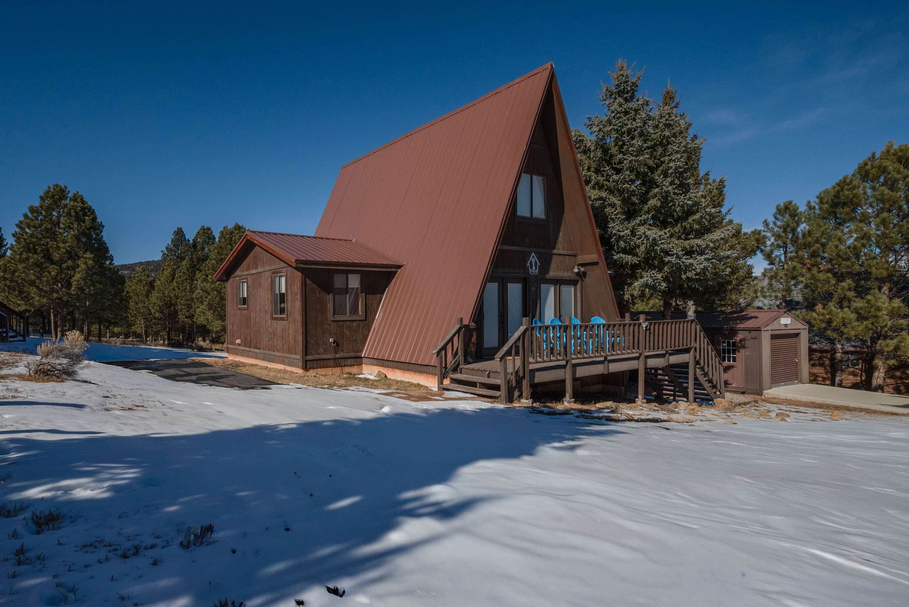 A two-story wooden cabin with a steeply pitched roof, a wooden deck, and a small porch with blue chairs.