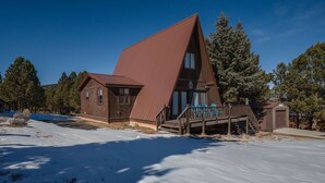 A two-story wooden cabin with a steeply pitched roof, a wooden deck, and a small porch with blue chairs. - The Ridge House Golf Skiing and Hiking (Angel Fire)