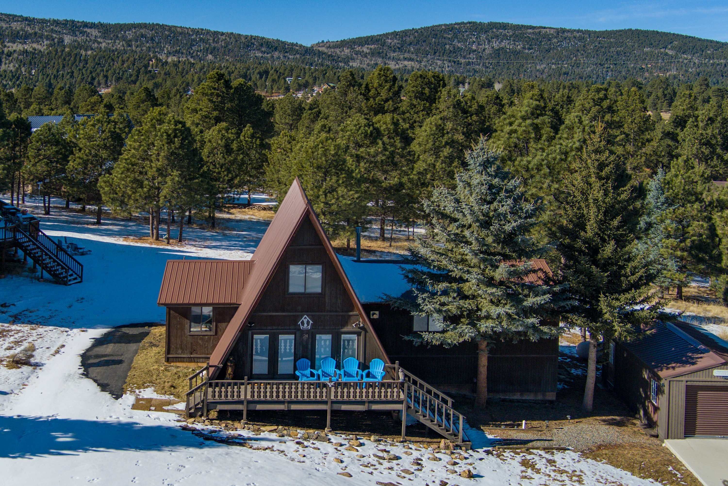 A cabin with a triangular roof, a porch with blue chairs, and a view of a snowy landscape with trees and hills in the background.