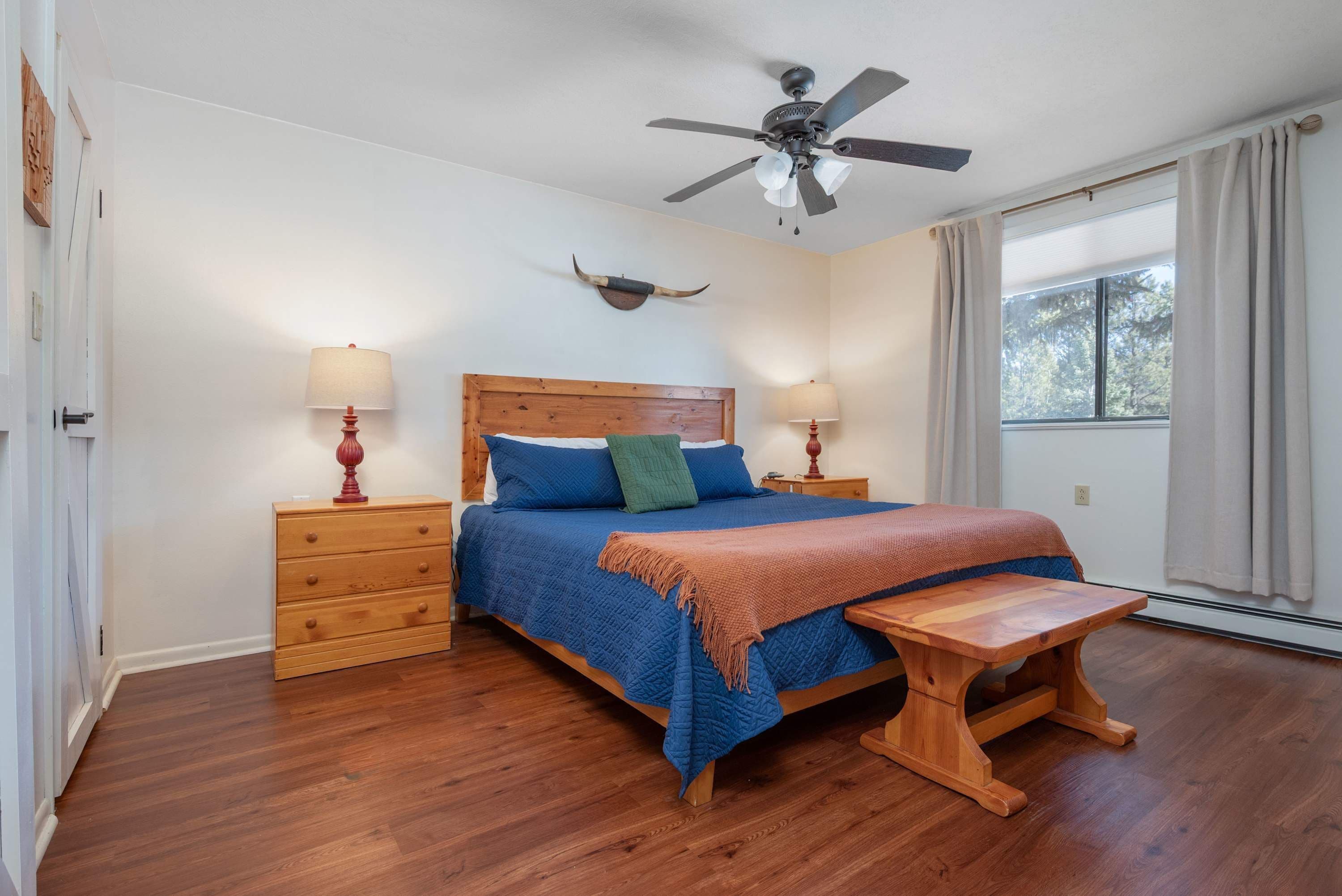 Un dormitorio con cama de madera, dos mesitas de noche con lámparas, un ventilador de techo, una ventana con cortinas y un banco de madera.