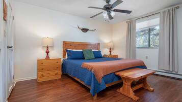 Un dormitorio con cama de madera, dos mesitas de noche con lámparas, un ventilador de techo, una ventana con cortinas y un banco de madera.