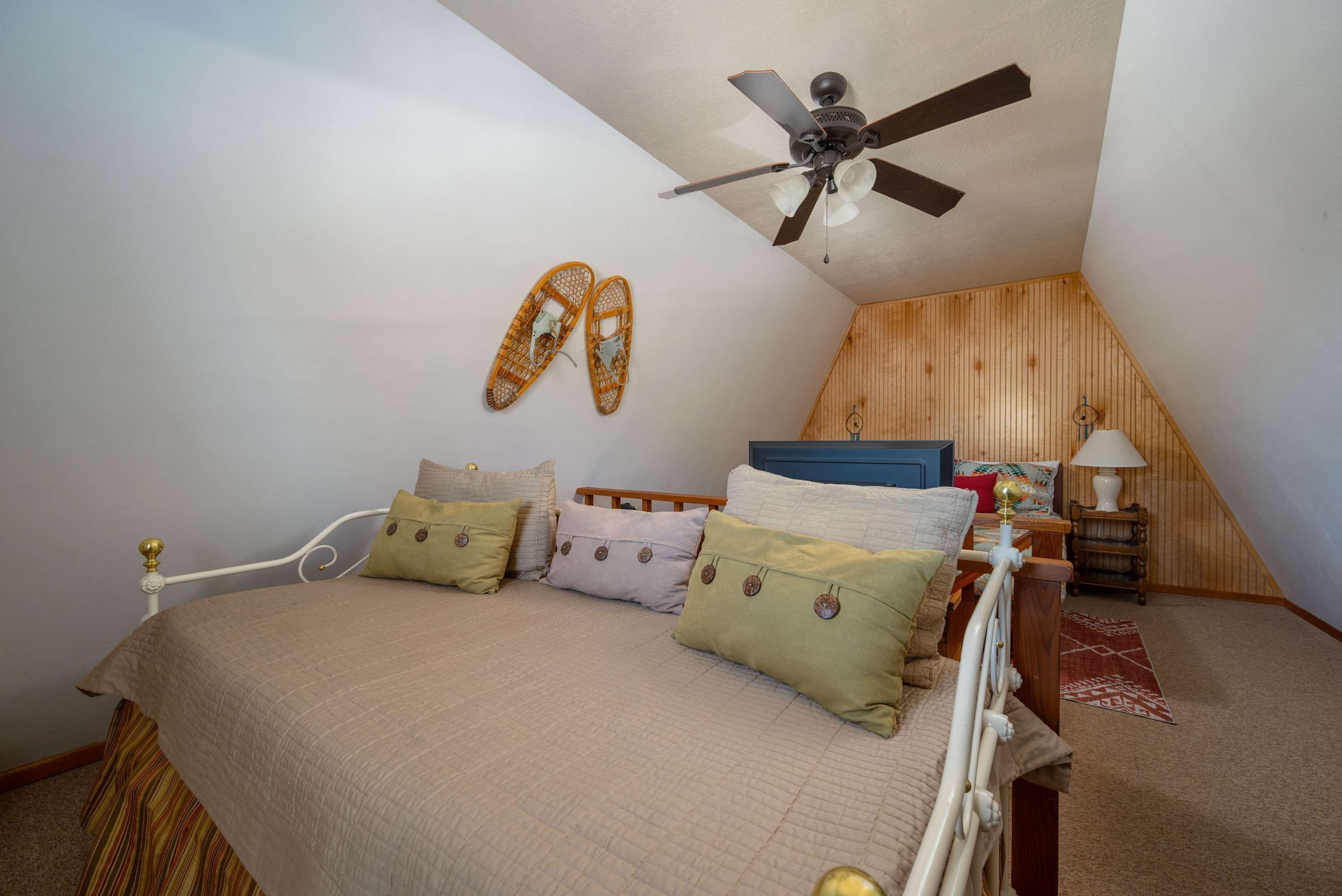 Une chambre à coucher avec un lit, des oreillers, un ventilateur de plafond, un plafond en pente en bois et une table de chevet avec une lampe.