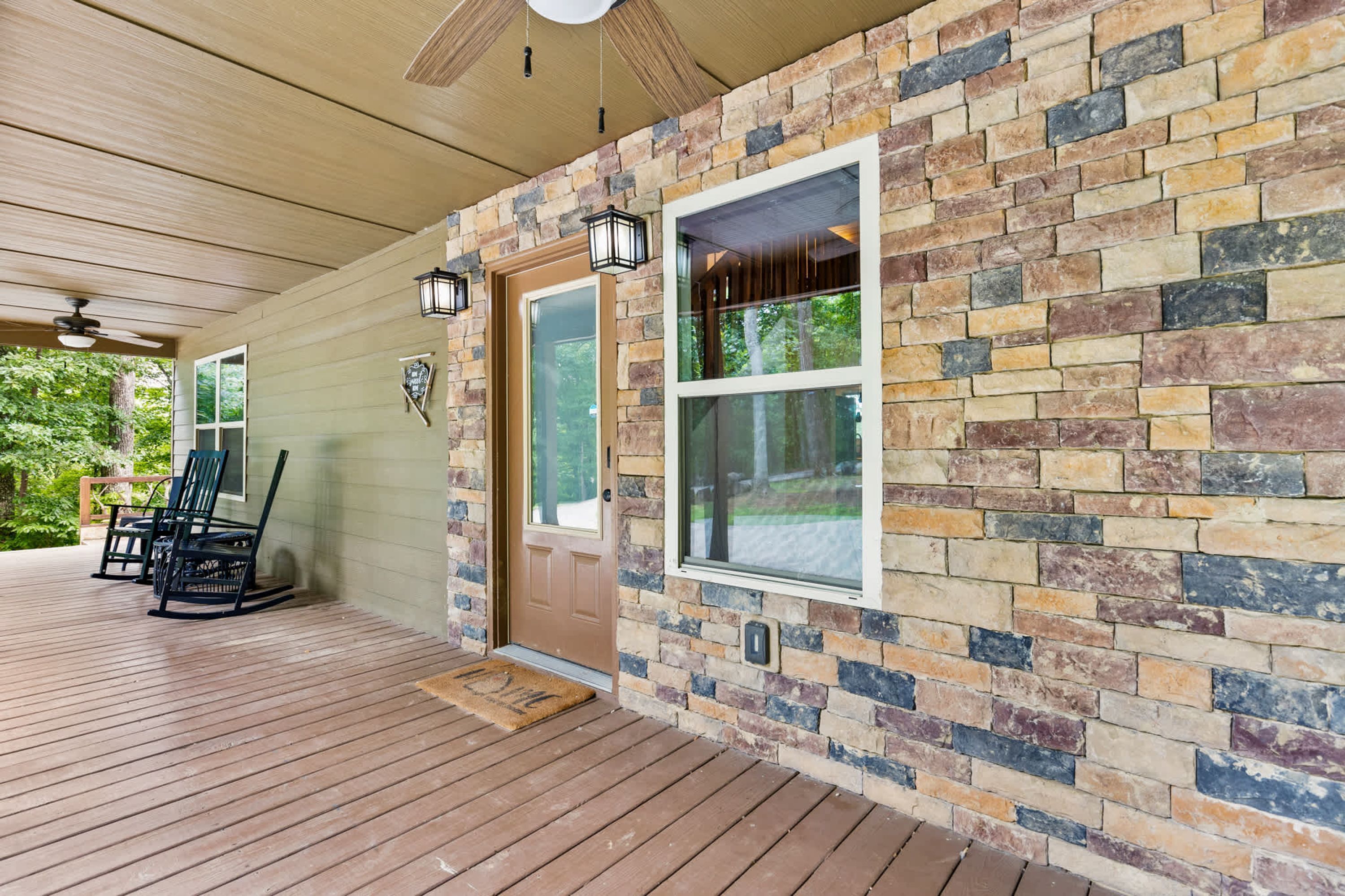 A covered porch with a wooden deck, a brown door, and a stone wall with a large window.