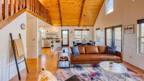 A spacious living room with a wooden staircase, a brown leather sofa, and a dining area with a view. - Valley Retreat Sleeps 10 Hot Tub Fire Pit (Blue Ridge)